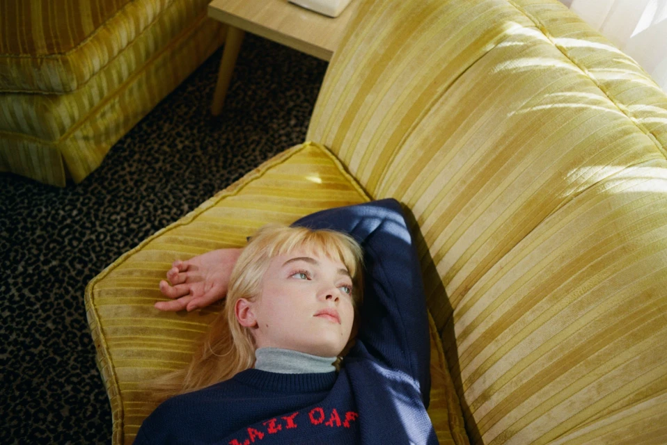 Young woman with blond hair lies on a yellow striped sofa, wearing a navy blue sweater with red letters. Animal print carpet below.
