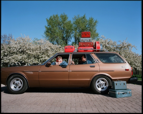 Vintage brown station wagon packed with luggage, driven by a woman, passenger, with additional suitcases stacked on the roof.