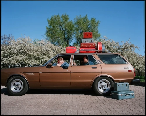Vintage brown station wagon packed with luggage, driven by a woman, passenger, with additional suitcases stacked on the roof.