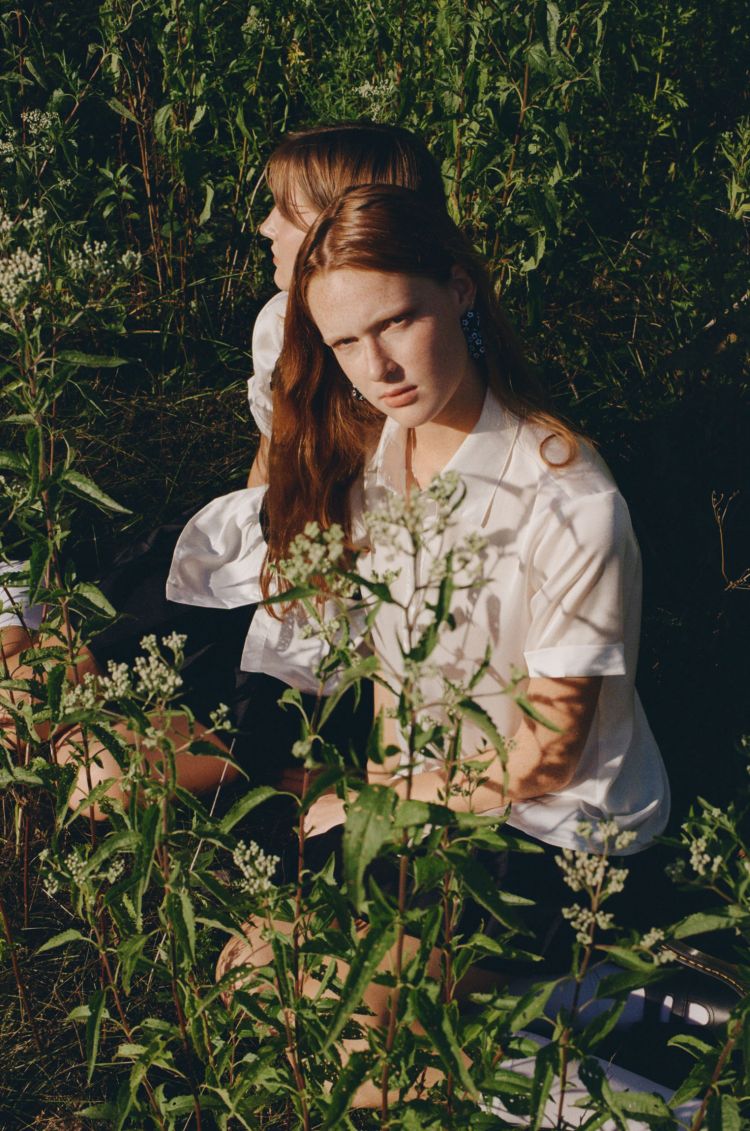 Two young women sit amongst tall, green foliage in an outdoor setting, bathed in sunlight. They both wear white tops and dark bottoms.