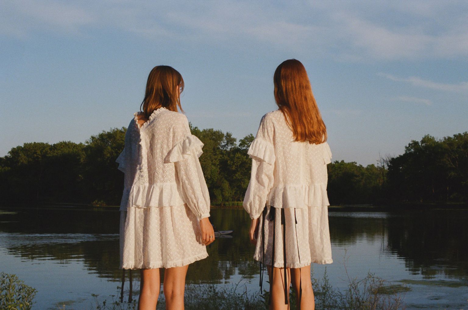 Two girls in white dresses stand by a lake surrounded by trees under a blue sky, creating a serene and youthful outdoor fashion scene.