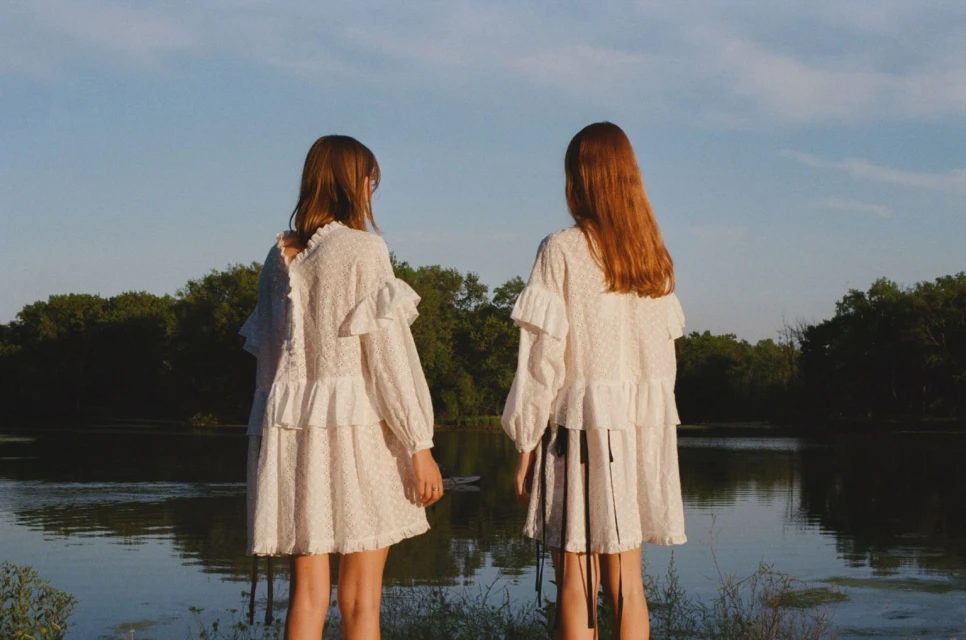 Two girls in white dresses stand by a lake surrounded by trees under a blue sky, creating a serene and youthful outdoor fashion scene.