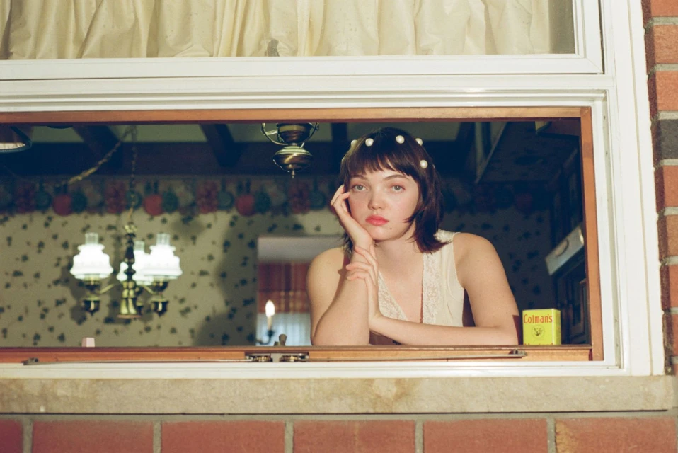 A young woman with bangs and pearl hair accessories gazes out of a window with red brick trim in an indoor setting with vintage wallpaper.