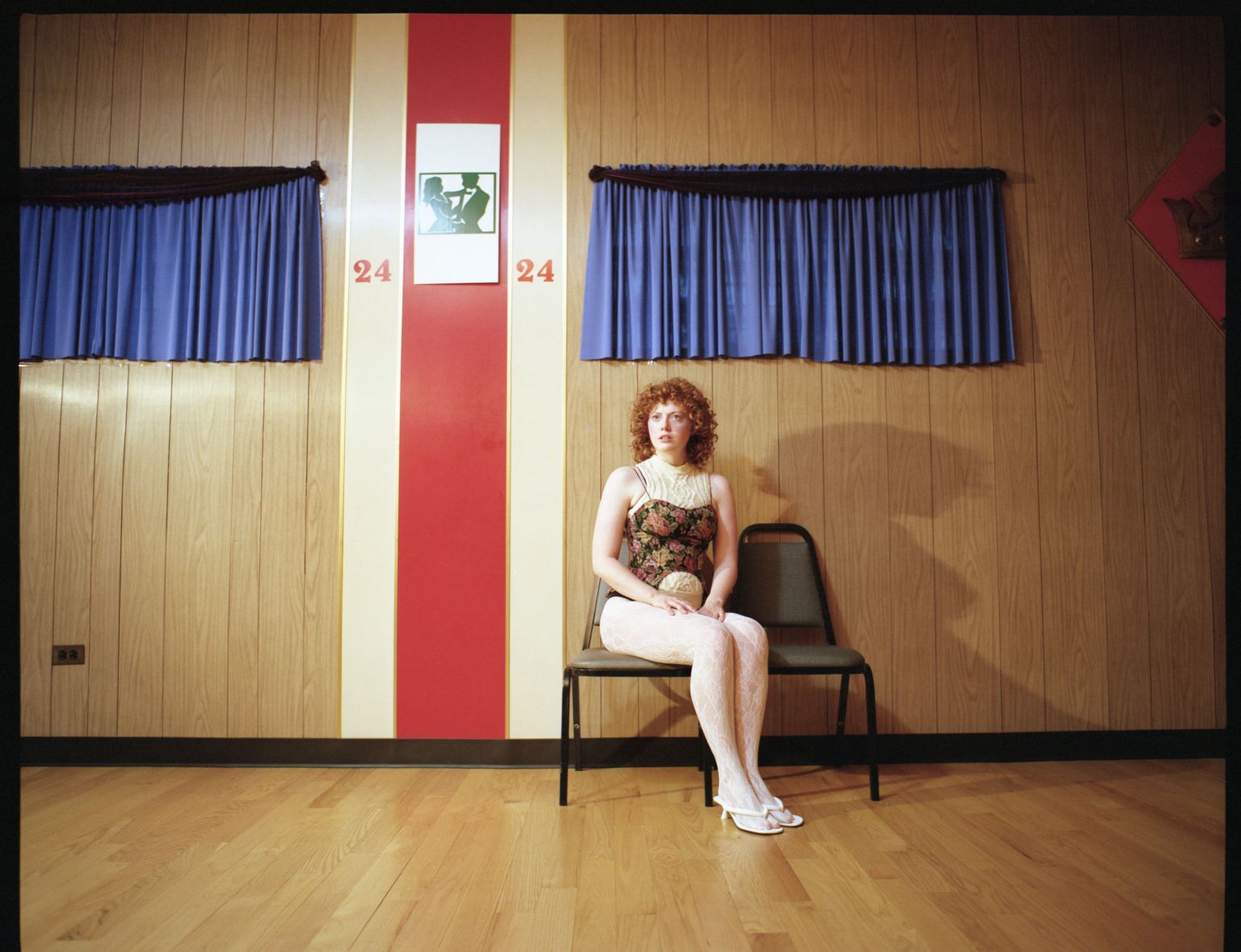 Red-haired woman sits formally in a waiting room with wood paneled walls and blue curtains, illuminated by a warm, theatrical lighting.