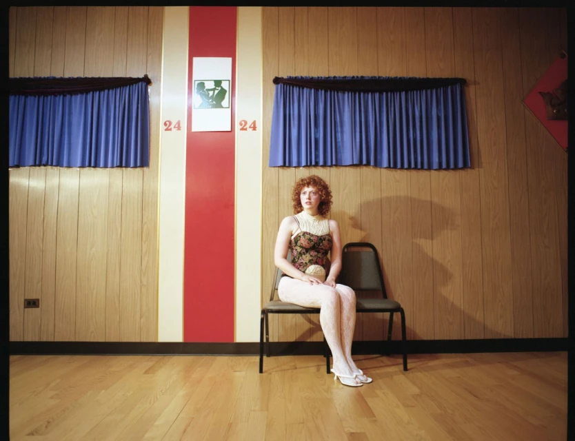 Red-haired woman sits formally in a waiting room with wood paneled walls and blue curtains, illuminated by a warm, theatrical lighting.