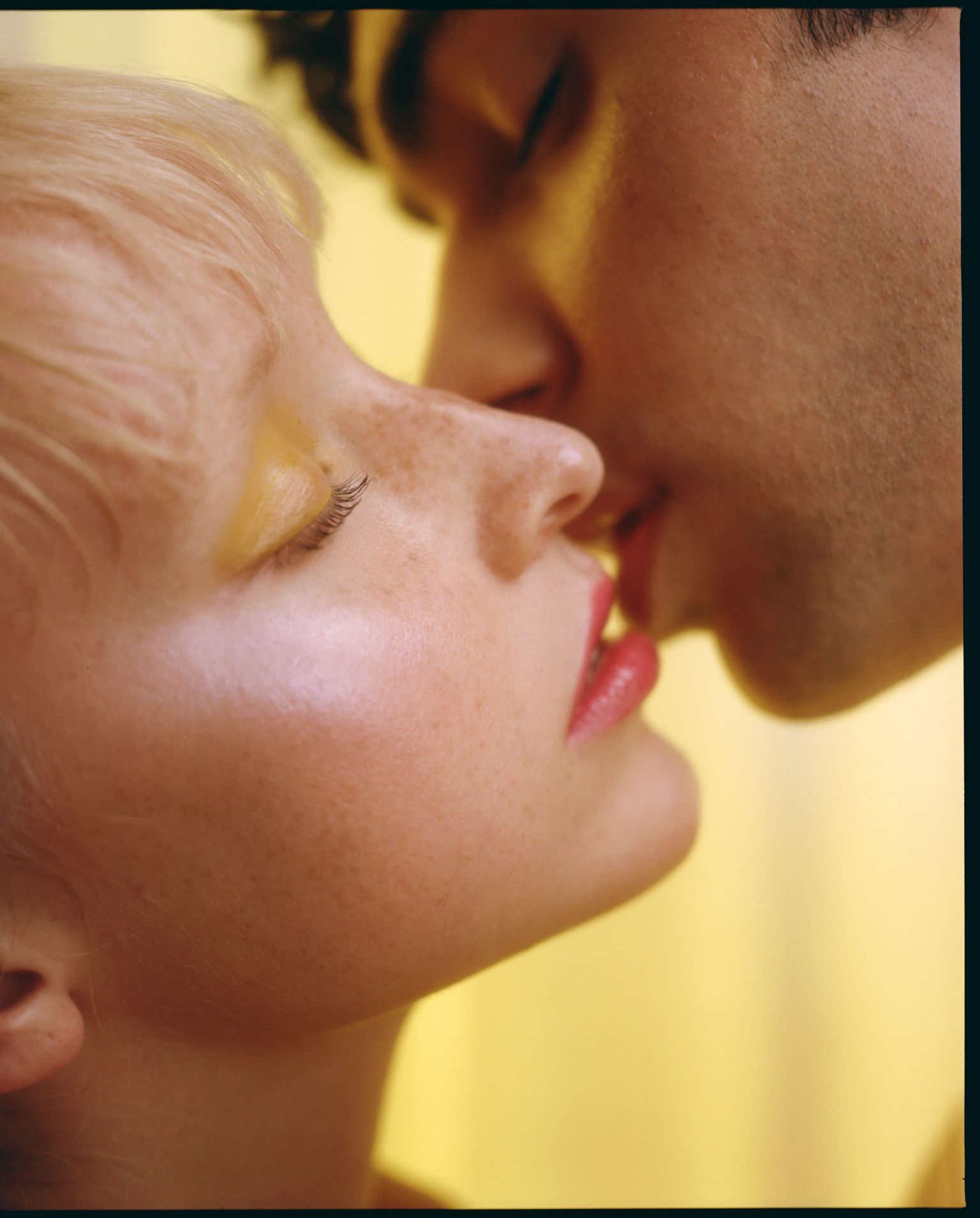 Close-up portrait of a couple kissing, woman with short blonde hair and yellow eye shadow, man with dark hair, on yellow background.
