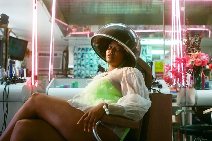 Woman with brown hair relaxing in hair salon chair under vintage dome hair dryer with pink neon lights, bright green top, and sheer sleeves.