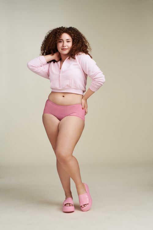 Smiling woman with curly hair poses in pink sweater and underwear against a neutral background in a studio setting.