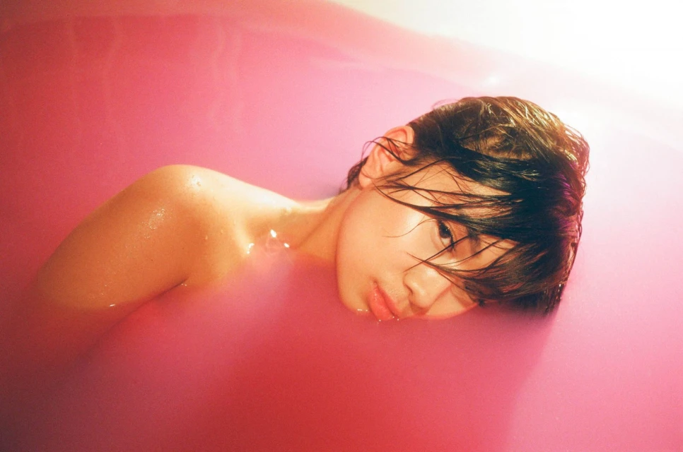 Woman with dark hair reclines in pink-tinted water in a bathtub, portrait-style, looking at the camera with a neutral facial expression.