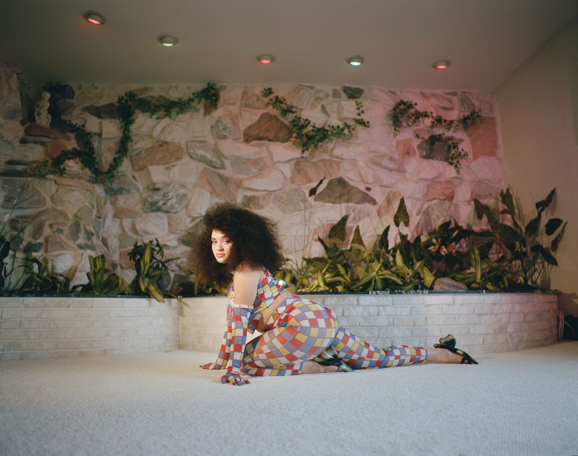 Woman in a patterned bodysuit poses on a carpeted floor in front of a stone wall with greenery, creating a vibrant and artistic composition.