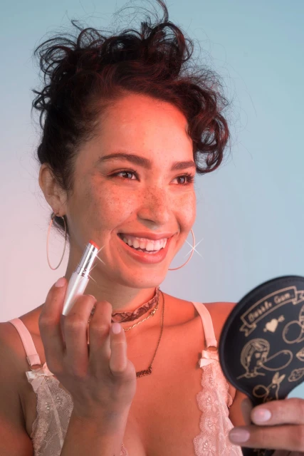 Smiling woman applies pink lipstick using a silver tube while looking in a hand mirror in a soft, flattering light setting.