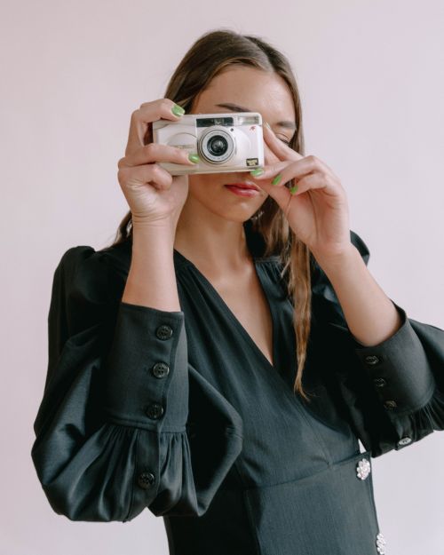Adult woman wearing black satin dress holds white digital camera up to eye for a photo in a light-filled indoor setting.