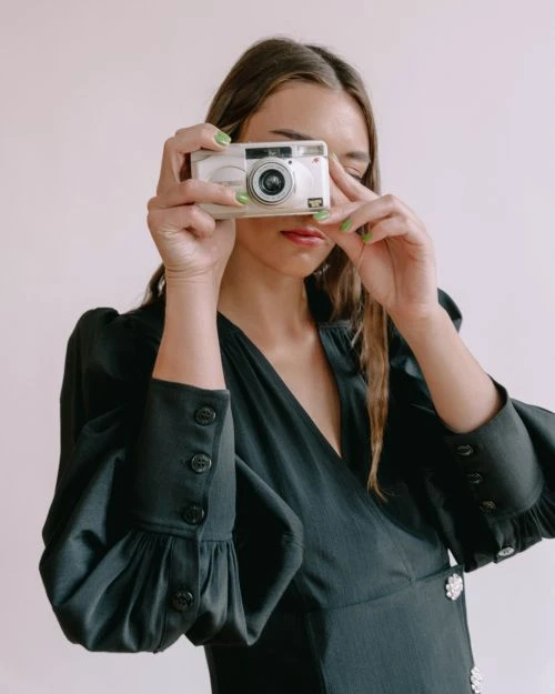 Adult woman wearing black satin dress holds white digital camera up to eye for a photo in a light-filled indoor setting.