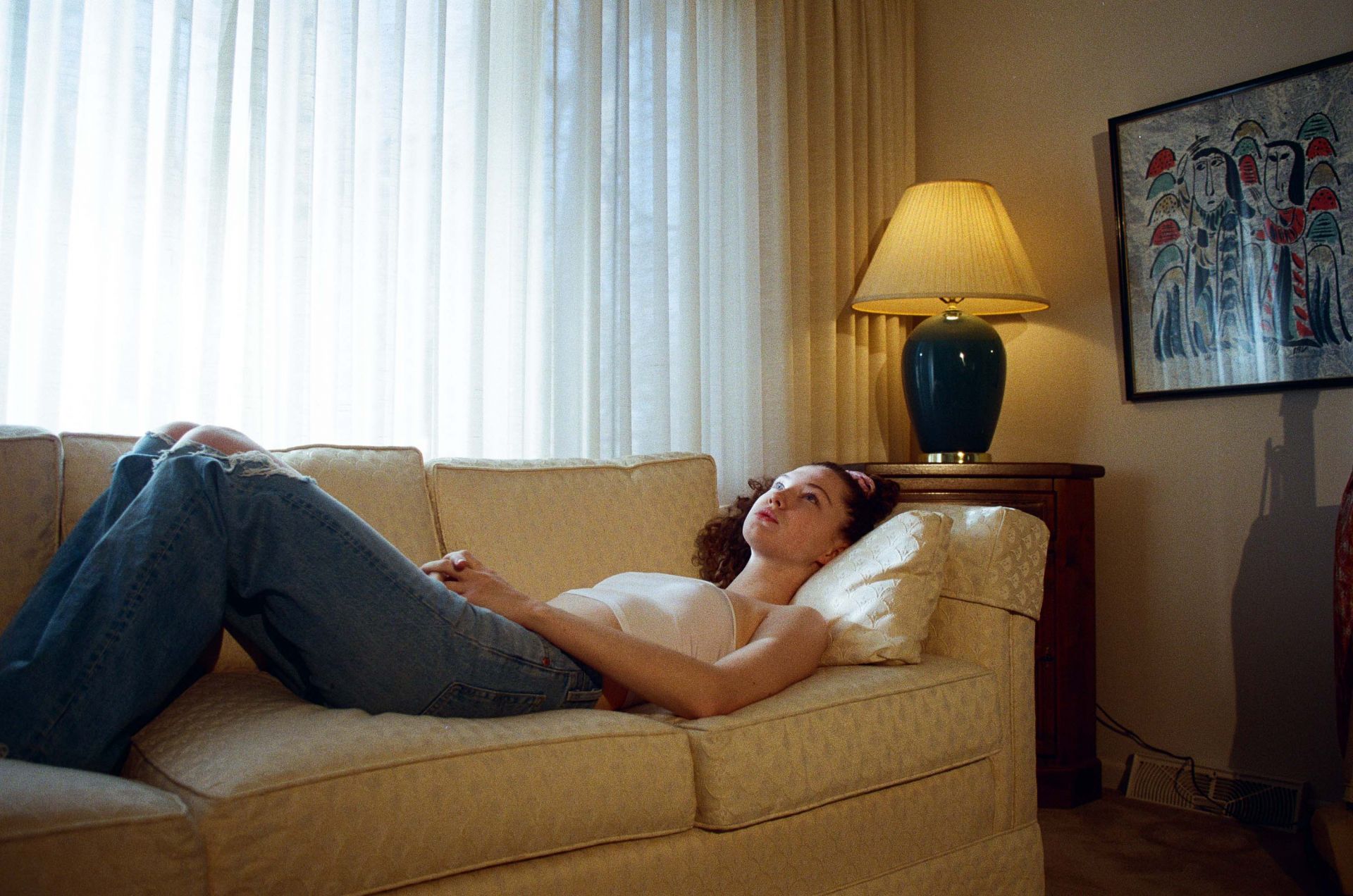 A young woman is relaxing on a tan couch, wearing a white tube top and blue jeans, in a brightly lit living room with a large window.