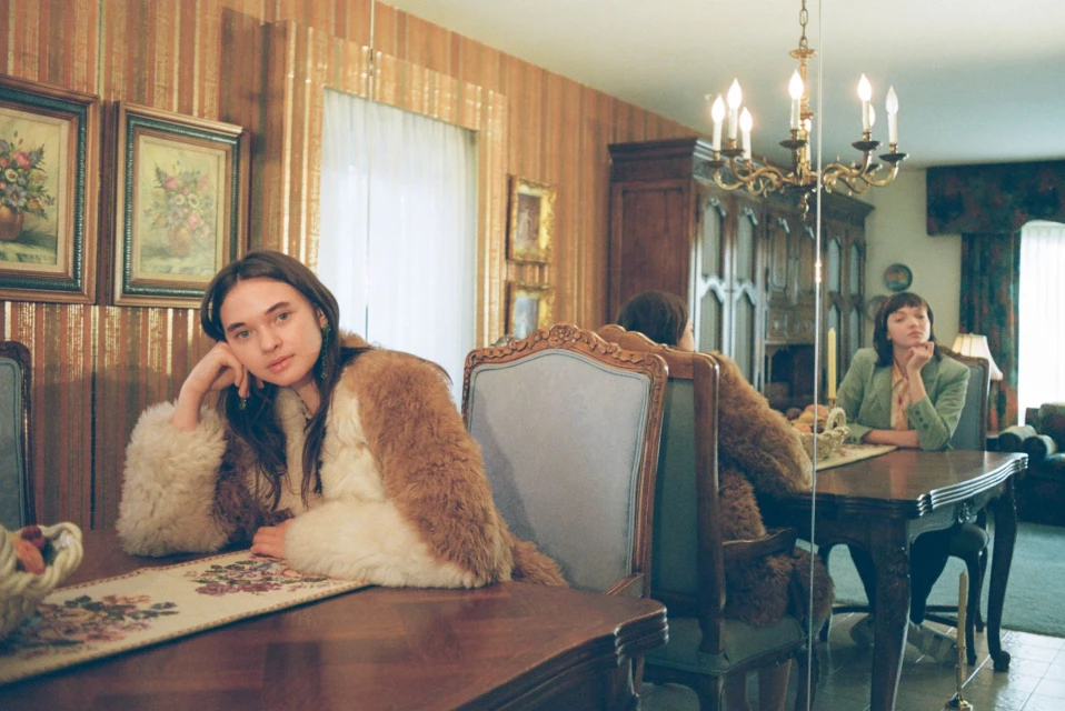Two women at a dining table, one woman in a fur coat in the foreground and her reflection in the background.