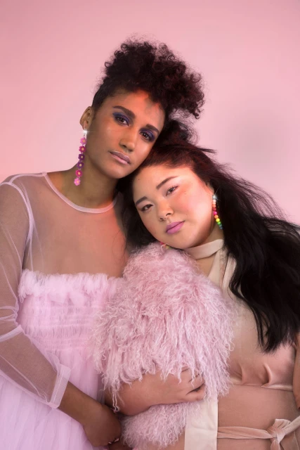 Two women in pink hues are posing together in a photo studio, wearing statement earrings and soft dresses. One woman has a fluffy pink coat.