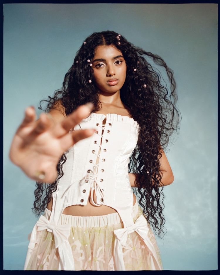 Portrait of a young woman with long, curly dark hair and fair skin, wearing a white corset top and a floral skirt with hand up.