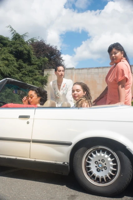Four young women ride in a white convertible under a partly cloudy sky, near trees and a wall, in a lifestyle fashion editorial image.