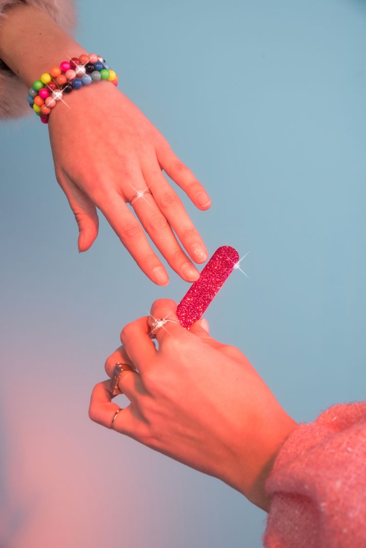 Two hands exchanging a glittery pink nail file against a light blue backdrop, accessorized with colorful beaded bracelets and rings.