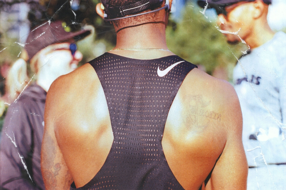 Back view of an athletic man wearing a black Nike tank top with two partially visible individuals in the background outdoors.