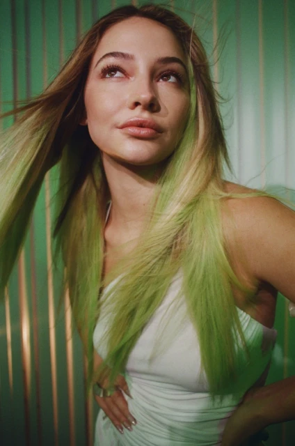 Portrait of a young woman with green-tipped blonde hair wearing a white dress, posing in front of a green and gold background.