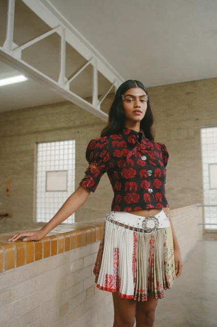 A young woman poses indoors wearing a black rose-patterned blouse, pleated patterned mini skirt, and a wide white belt with a silver buckle.