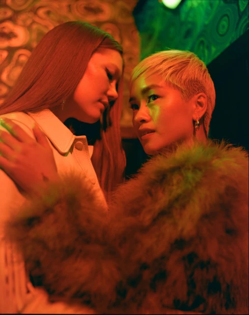 Two young women pose closely indoors with warm lighting. One with long brown hair and one with short blonde hair, wearing fashionable jackets.