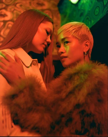 Two young women pose closely indoors with warm lighting. One with long brown hair and one with short blonde hair, wearing fashionable jackets.