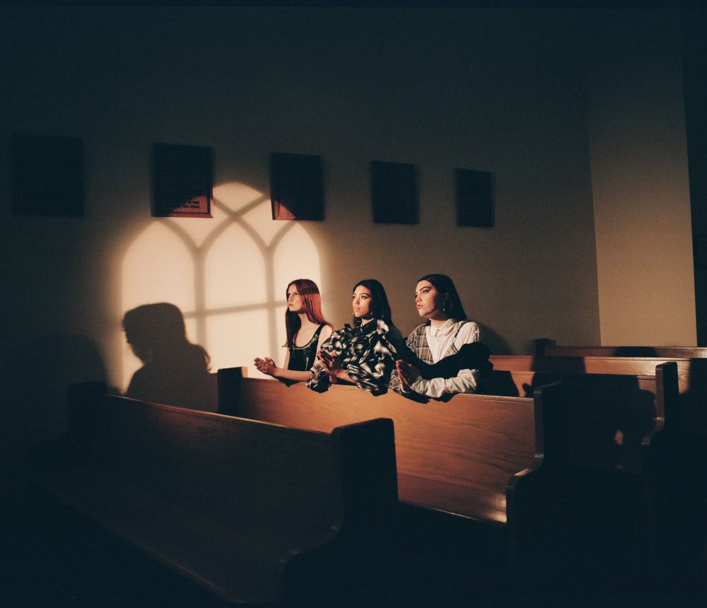 Three young women sit in a wooden church pew bathed in the light from a window, creating a serene and reflective atmosphere.
