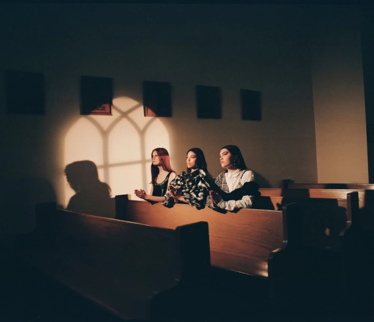 Three young women sit in a wooden church pew bathed in the light from a window, creating a serene and reflective atmosphere.