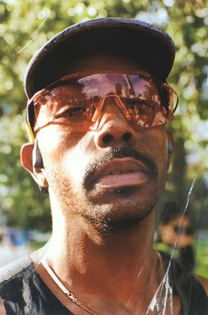 Portrait of a man wearing orange-tinted sunglasses, a black cap, and a dark tank top outdoors.