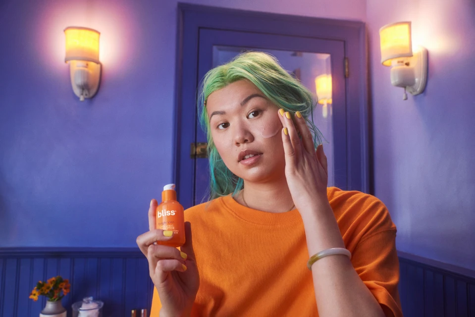 An Asian woman with green-blue hair is indoors applying skincare to her face, holding a bottle of bliss product, with a blue background.