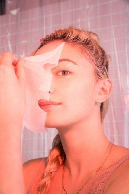 Adult female removing a sheet mask from her face in an indoor environment. The lighting is pink and shows the facial mask detail.