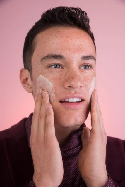 Close-up of a young adult male with freckles applying facial cream against a pink background, wearing a burgundy shirt and blazer.