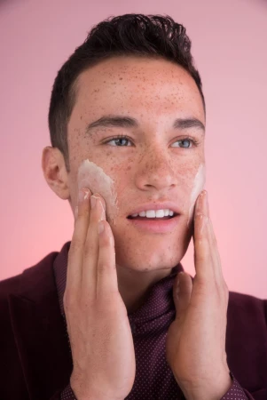 Close-up of a young adult male with freckles applying facial cream against a pink background, wearing a burgundy shirt and blazer.