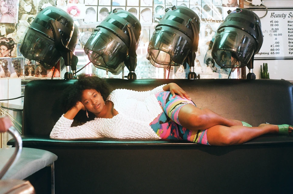 Woman lounges in a salon chair under vintage hair dryers. She wears colorful shorts and a white textured top in a beauty salon setting.