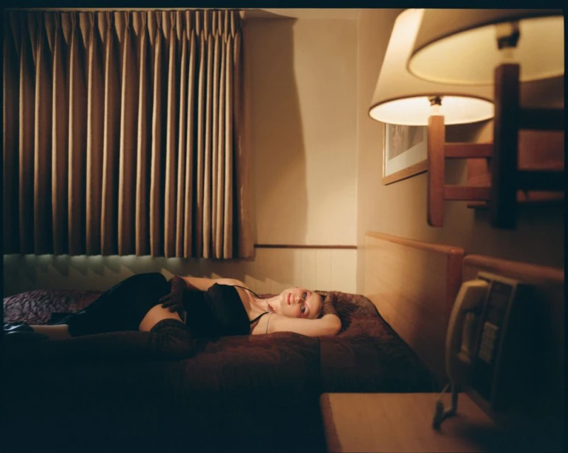 A woman in a black top lies on a bed in a softly lit room with curtains, a wall lamp, and a telephone. The scene evokes a calm mood.