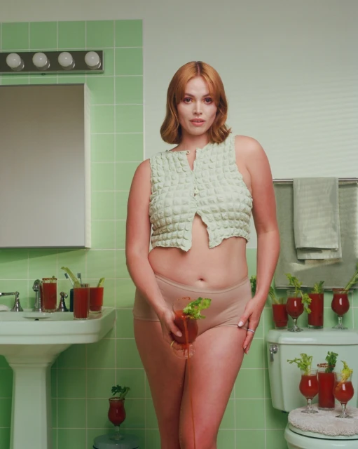 An adult woman poses in a bathroom wearing a light green cropped top and underwear, while holding a Bloody Mary cocktail.