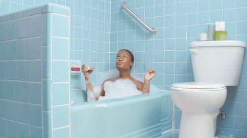 Woman relaxes in a bubble bath with a glass of red wine in a bright blue tiled bathroom with a toilet, creating a tranquil scene.