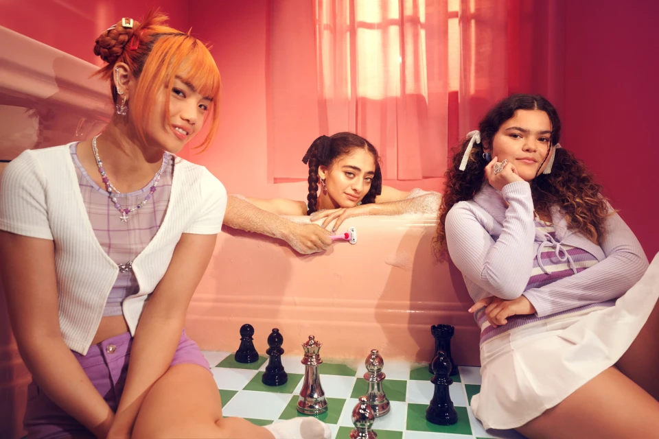 Three diverse young women are posing indoors with a bathtub and a chess set, dressed in casual coordinated pastel fashions in a pink room.