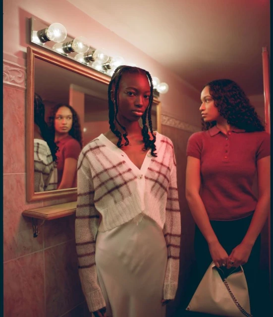 Two young women pose in an indoor bathroom setting with vintage lighting and patterned wall tiles, reflected in a mirror.