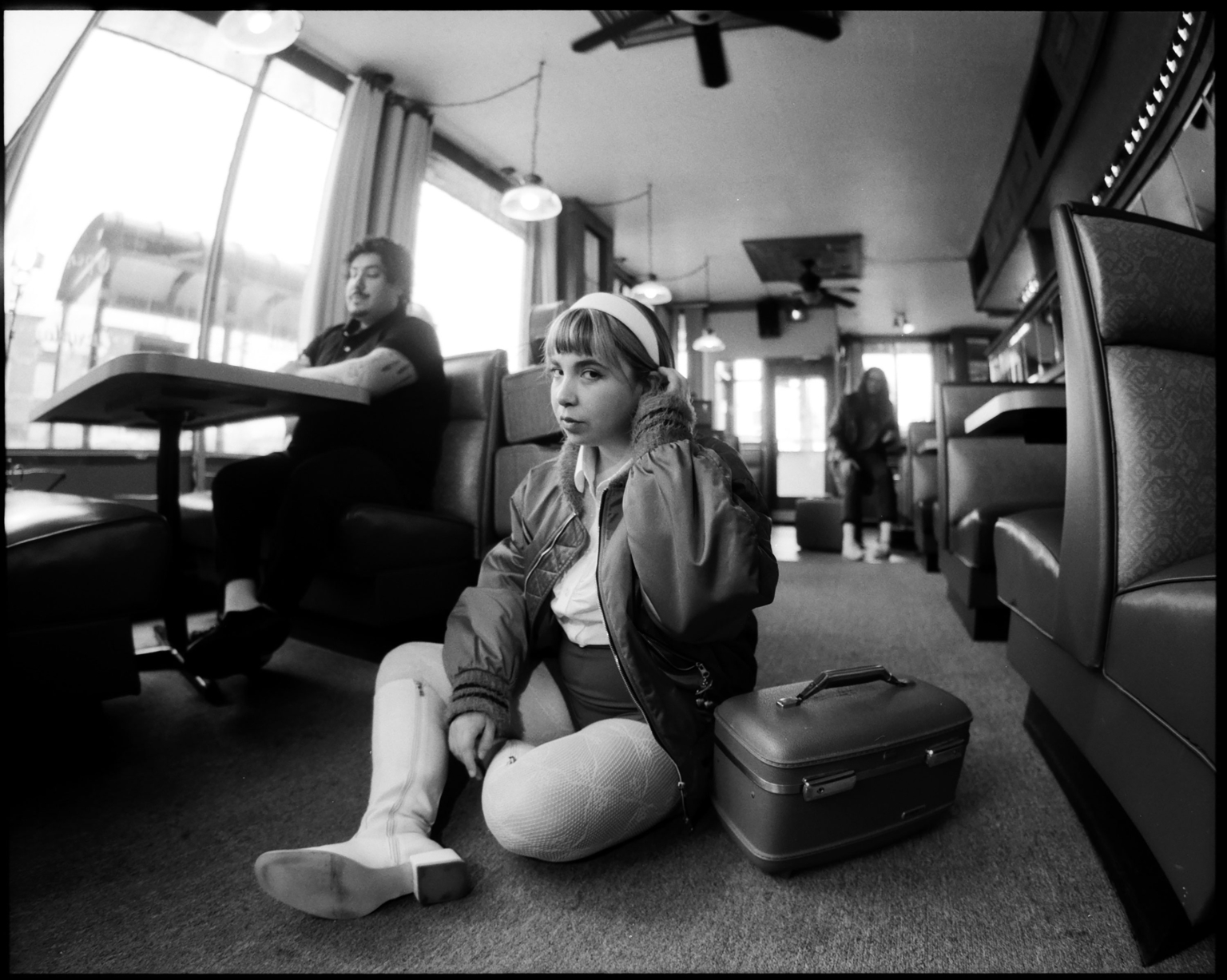 Black and white photo in a diner featuring a woman seated on the floor with a suitcase and a man in a booth in the background.