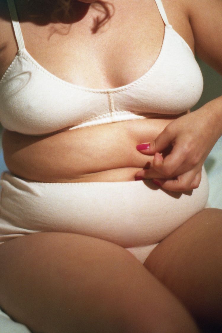Close-up of a person pinching the skin on their abdomen, wearing a white bra and underwear, with pink nail polish visible.