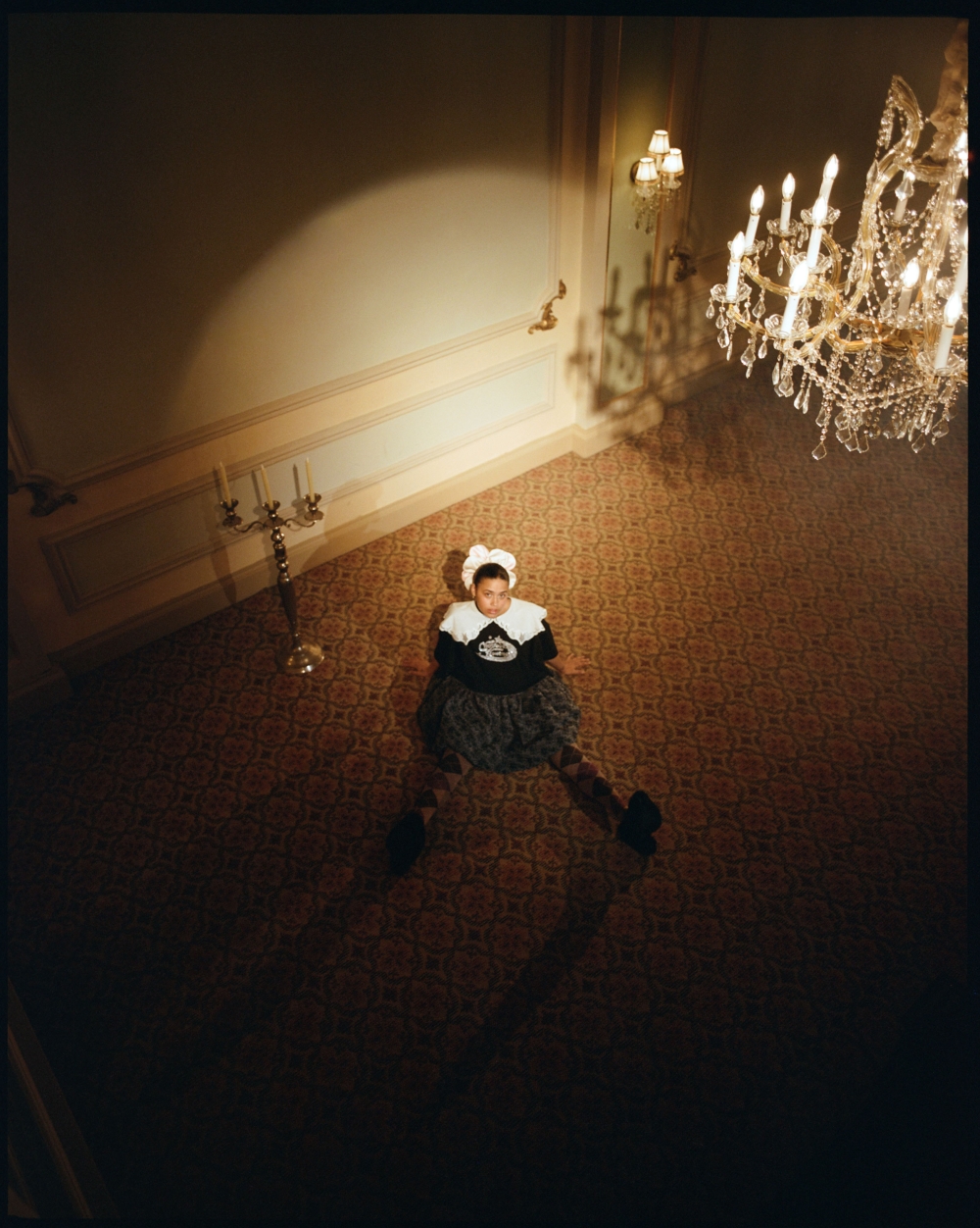 Overhead shot of a woman in a decorative dress sitting on a patterned floor in an elegant foyer with a chandelier.
