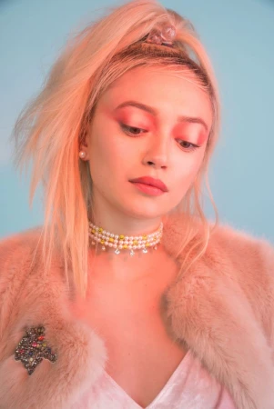 Close-up portrait of a woman with blonde hair in a high ponytail wearing a pearl choker and pink faux fur jacket against a blue background.