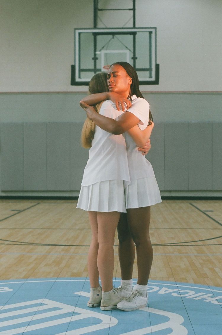 Two women in white athletic outfits embrace on a blue basketball court. Focus on teamwork, friendship, and sportsmanship.