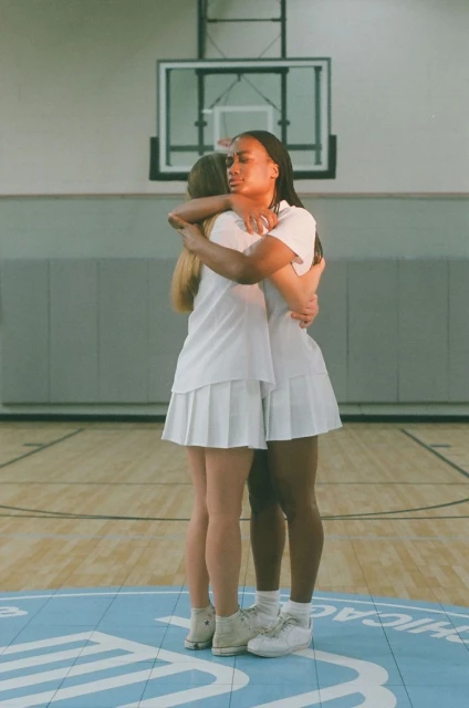 Two women in white athletic outfits embrace on a blue basketball court. Focus on teamwork, friendship, and sportsmanship.