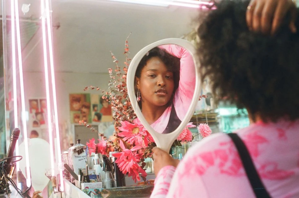 A young adult woman looks into an oval-shaped hand mirror with a white handle, posing in an indoor dressing-room-like setting with pink floral elements.