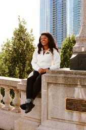 Smiling black woman sitting on a stone wall wearing a white zip-up jacket and black leggings outdoors in Los Angeles.