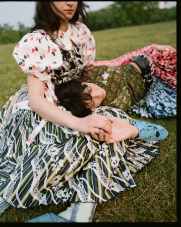 Two people resting in a grassy park, one sitting upright in a patterned dress while the other lies with their head on their lap.